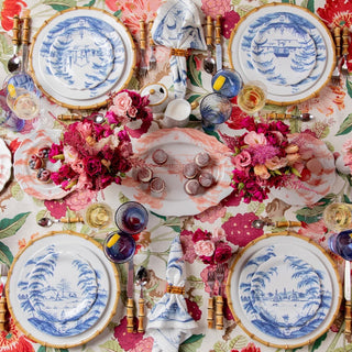 A styled tabletop setting with four blue-and-white porcelain plates featuring tropical landscape illustrations, gold-rimmed edges, matching napkins, blue and amber glasses, and floral centerpieces on a pink-and-white patterned tablecloth.